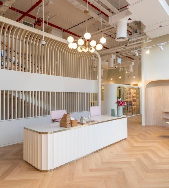 A modern reception area with a sleek, curved reception desk at the center, decorated with wicker baskets and a vase containing small decorative objects. The background features an arched structure with vertical wooden slats that house the sign 'the bureau.' To the right, a cozy seating area includes two white armchairs, a small round table, and potted plants. The ceiling is adorned with industrial-style exposed pipes and ducts, with hanging lights providing illumination. The floor is designed with a herringbone pattern.